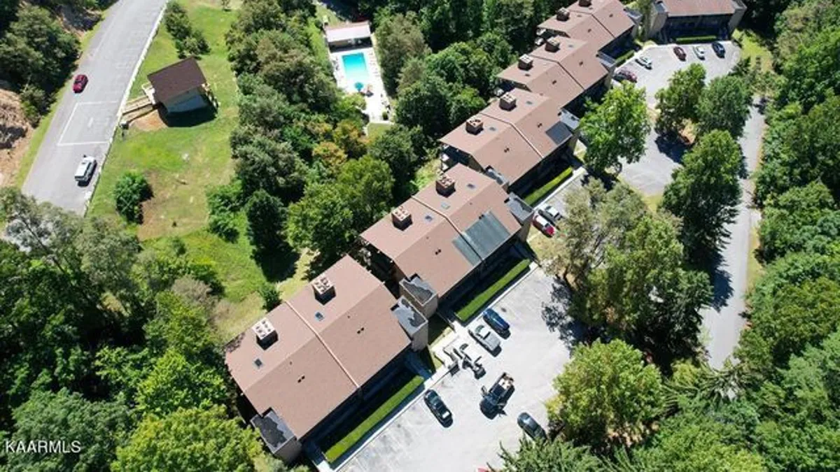 A wide view of the community shows rows of townhouses, a sparkling swimming pool, surrounding trees, and a parking area. The photo captures a peaceful, community-oriented mood, with ample space for relaxation, socializing, and outdoor activities.
