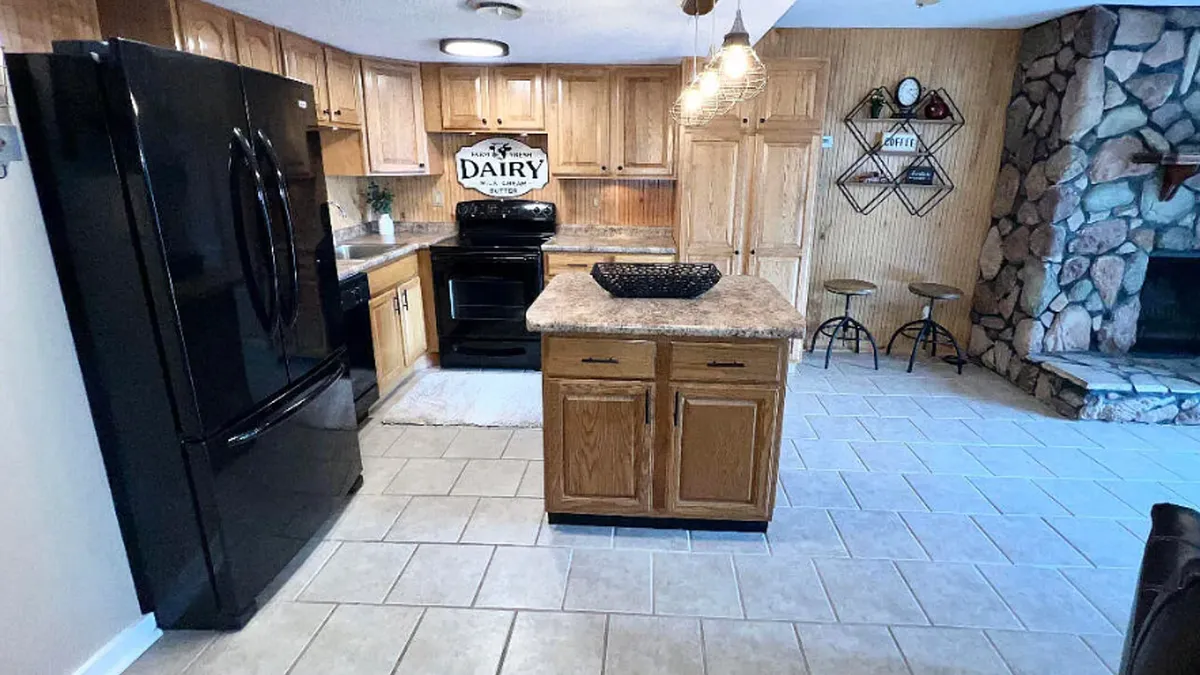 A seamless open-concept space showcasing a kitchen with black appliances, wooden cabinetry, a kitchen island, and a stone fireplace just beyond. The area is designed for both cooking and socializing, with the layout fostering togetherness and warmth.