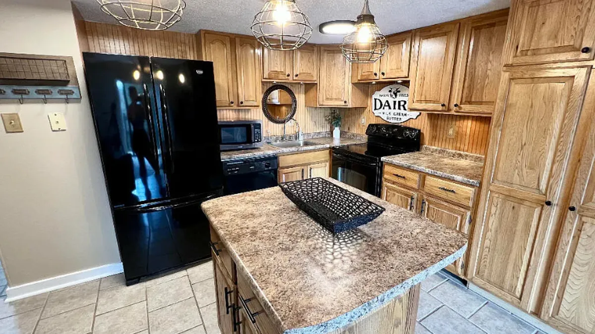 A kitchen-focused view with a central island topped by a decorative basket, surrounded by black appliances and warm wood cabinetry. Pendant lighting adds a welcoming touch, and the island provides ample space for food prep or casual conversation.