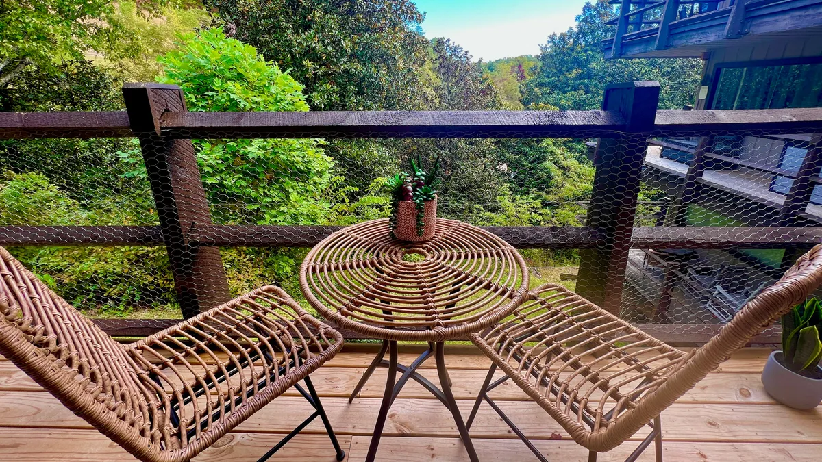 A cozy outdoor scene featuring a round wicker table with two matching chairs set on a balcony or patio. Potted plants add a homey touch, and lush green trees create a serene, natural backdrop. The mood is peaceful and intimate, perfect for enjoying coffee or quiet conversation while taking in the mountain air.
