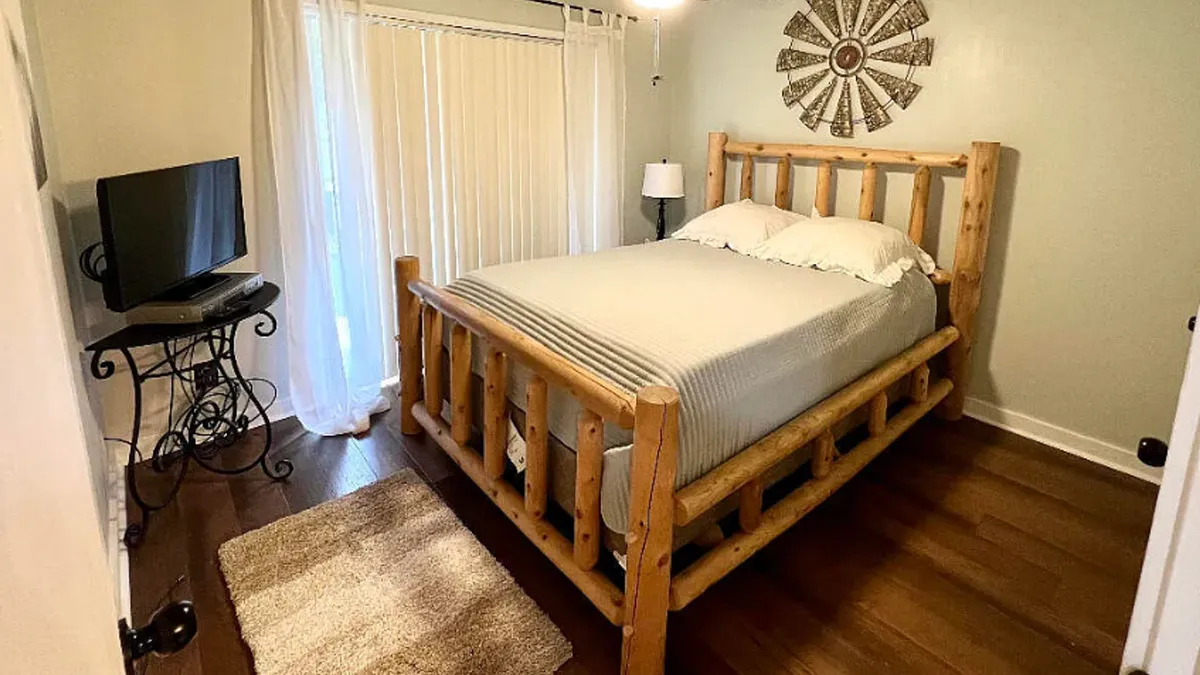 A cozy master bedroom featuring a log bed dressed in fresh linens, a TV stand with television, a large window with curtains that lets in natural light, and tasteful wall decor. The overall atmosphere is calm and inviting, perfect for relaxation.