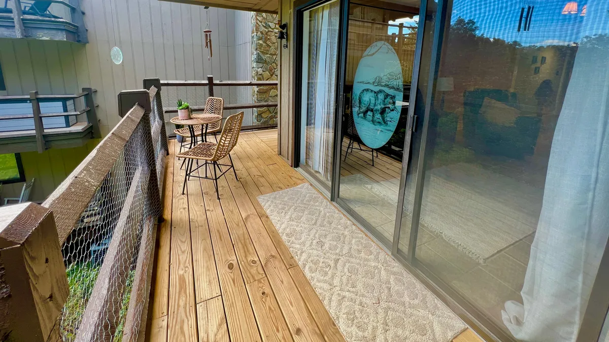 A charming wooden deck with a bistro table and chairs, a decorative wind chime, and a large glass door leading inside. The space feels private and invites guests to relax with a beverage while taking in the serene wooded views or sunsets. The mood is both inviting and tranquil, perfect for unwinding together.