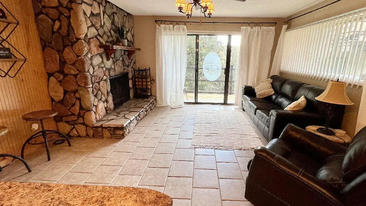 A warm and inviting interior featuring a stone fireplace as a focal point, large windows framing scenic woodland views, and plush, comfortable seating arranged to encourage relaxation and conversation. Natural light fills the room, and the rustic decor enhances the cozy, mountain cabin feel.