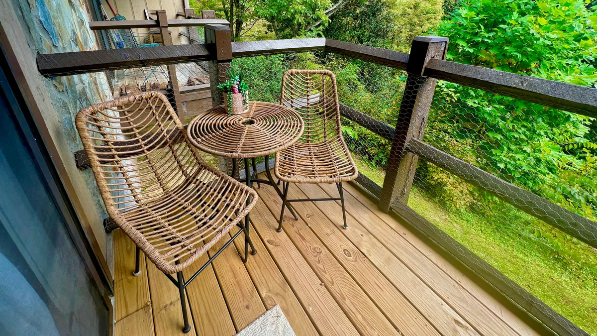 A cozy patio scene with rattan chairs and a round table, set against leafy greenery. The area is suitable for enjoying morning coffee, reading, or quiet moments alone.