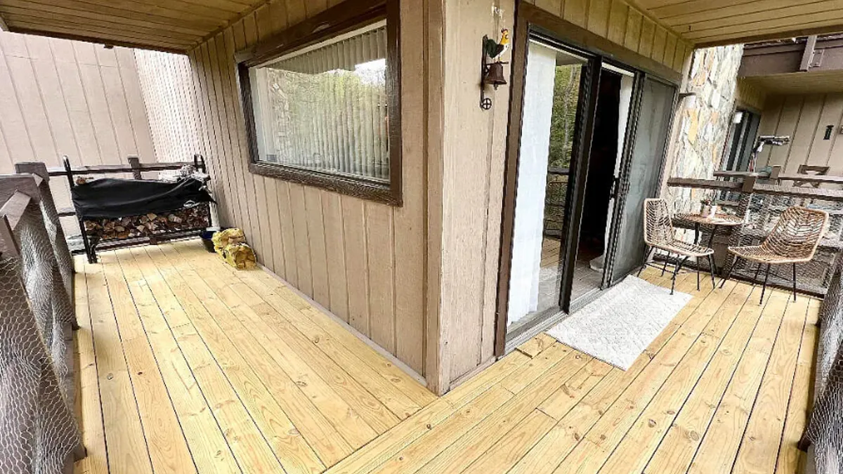 This image shows a wooden deck with comfortable outdoor seating and firewood storage, set against a backdrop of lush greenery. The deck is well-appointed for relaxation, with morning sunlight spilling onto the space—suggesting ease of access from parking to the welcome area.